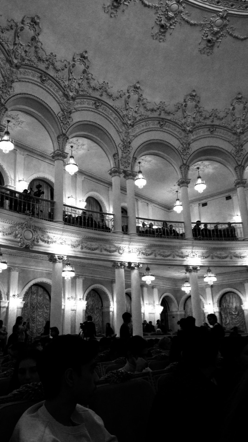 A grand theatre auditorium with ornate balconies and chandeliers and an audience gathering below, in black and white
